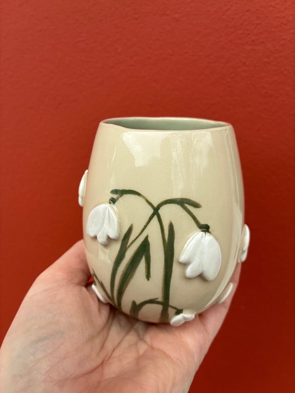 Hånd holder en beige keramikvase med hvide snepryd blomster og grønne stilke foran en rød væg.