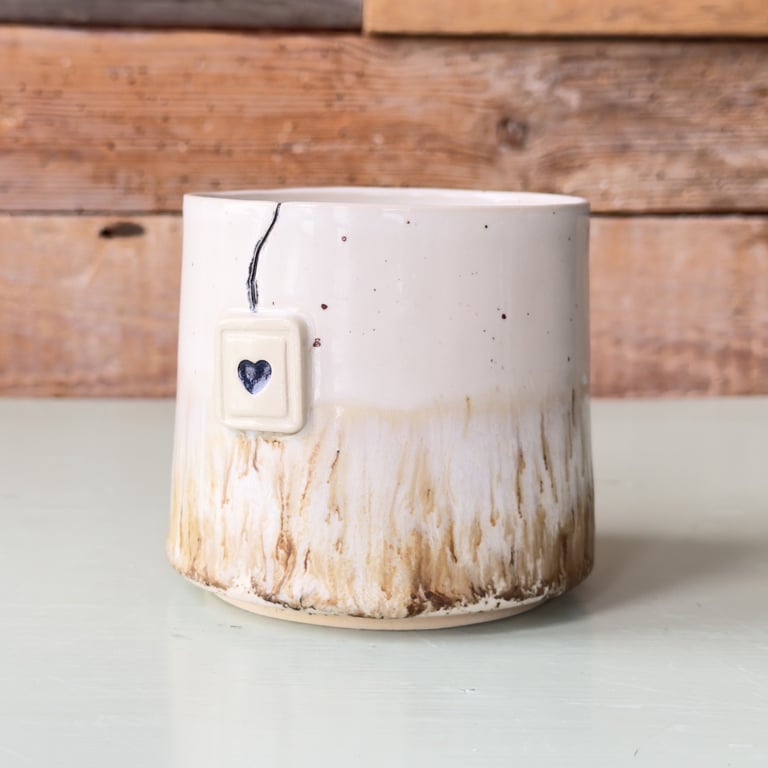 Keramikkop med beige-brun glasur og et lille firkantet vedhæng med et hjerte, placeret på en lys overflade med en trævæg i baggrunden.