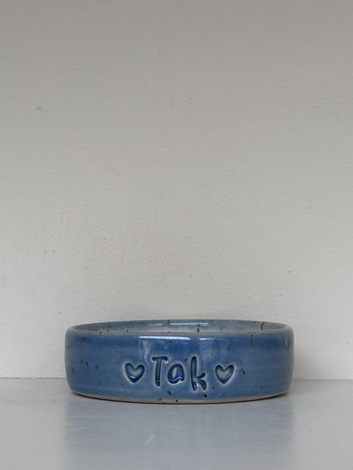 Blå keramikskål med teksten "Tak" flankeret af to hjerter, placeret på en hvid overflade mod en beige væg. (1)