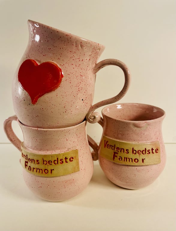 Tre lyserøde keramikkrus med hank og tap, to med teksten "Verdens bedste Farmor" og et med et rødt hjerte.