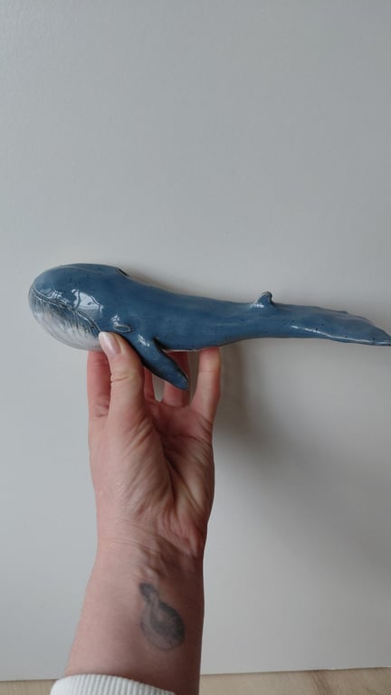 Hånd der holder en blå hvalfigur i keramik mod en lysegrå baggrund.