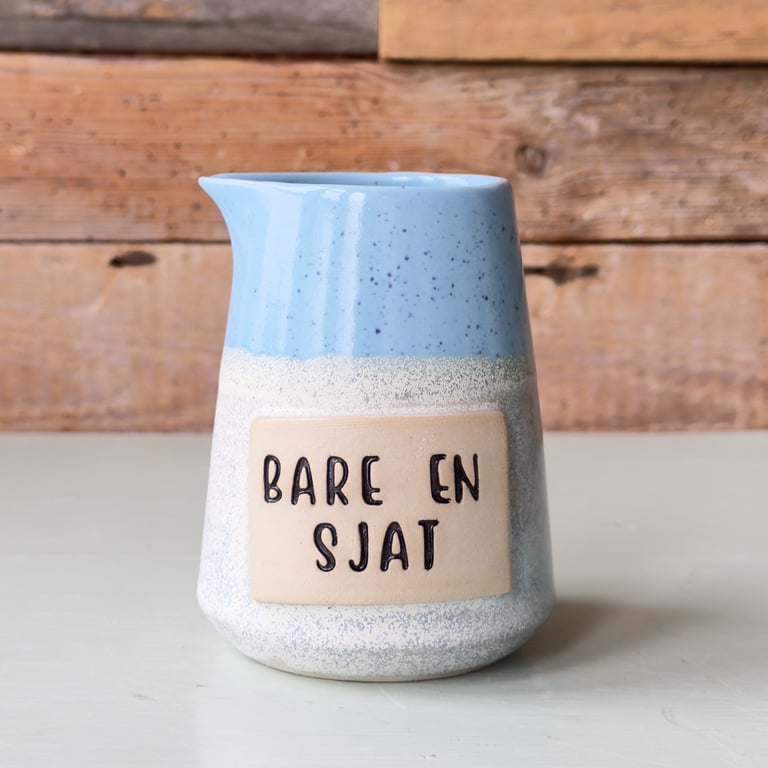 Keramisk kande med blå og beige glasur samt teksten "Bare en sjat" på forsiden, placeret på en lys overflade med en træbaggrund.