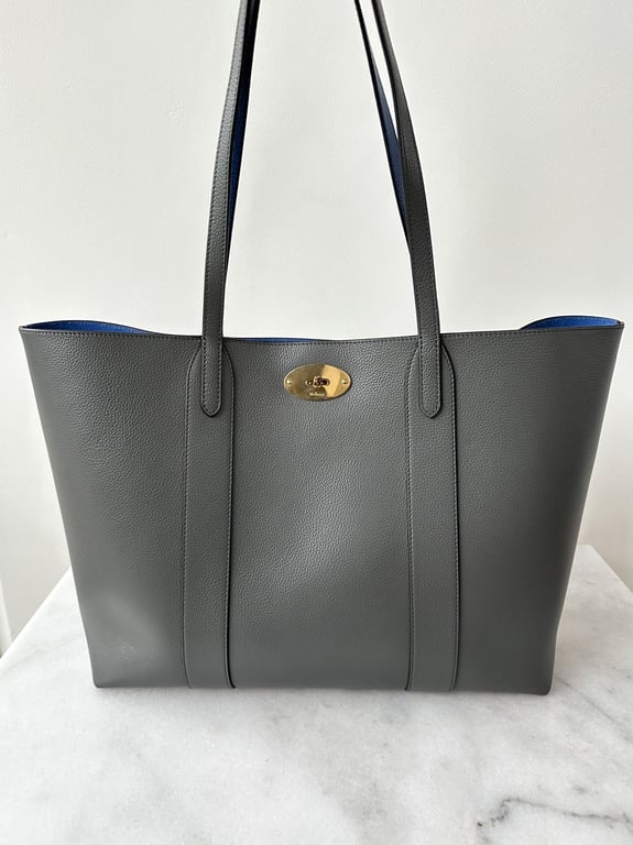 Grå læder shopper-taske med to skulderremme og gyldent spænde foran, stående på en hvid marmorflade.