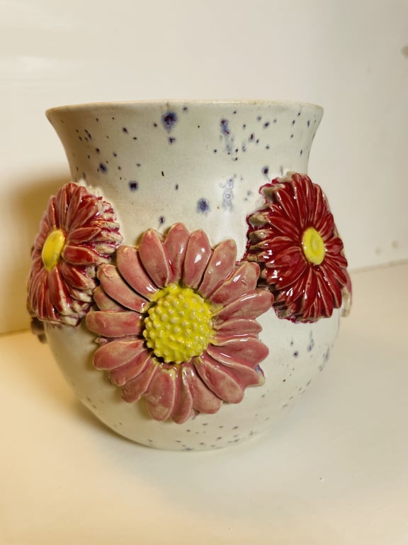 Keramisk, hvid vase med blå pletter og tre store reliefblomster i røde og rosa nuancer med gule midterdele.