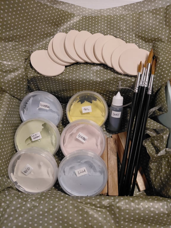 Malergrej i en kasse med små malerbørster, runde træskiver og flere plastikbeholdere med maling i forskellige farver mærket "lyseblå", "gul", "sort", "lysegrøn", "lyserød", "hvid" og "lilla".