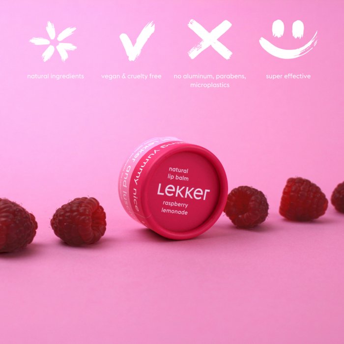 En pink læbepomade med smag af hindbær og lemonade omgivet af friske hindbær på en lyserød baggrund. Over produktet er hvide ikoner med tekster, der fremhæver naturlige ingredienser, vegansk og cruelty free, fri for aluminium, parabener og mikroplas