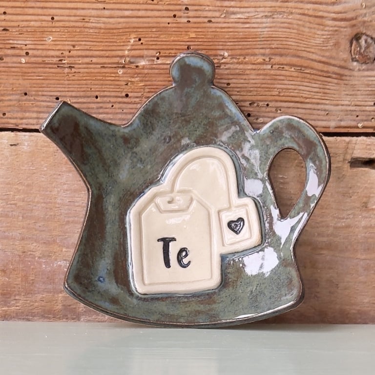 Keramisk teblad-holder formet som en tekande med en tebrevsdekoration og teksten "Te" på forsiden, placeret foran en rustik trægavl.