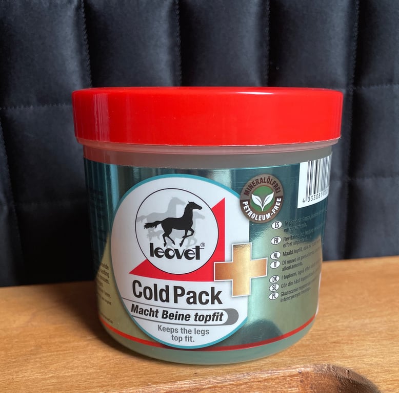 Beholder med Leovet Cold Pack, en salve til at holde benene i form, med rødt låg og grøn etikette, stående på et træbord foran en mørk baggrund.