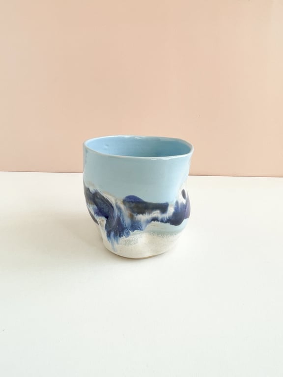 Lys blå keramikkrus med bølgende, abstrakt mørkeblå og hvide mønstre, placeret på en hvid overflade mod en beige baggrund.