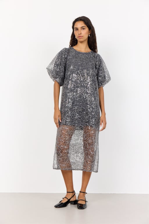 Kvinde iført gennemsigtig, sølvfarvet kjole med kropsnært underkjole og puffede ærmer, stående mod hvid baggrund.