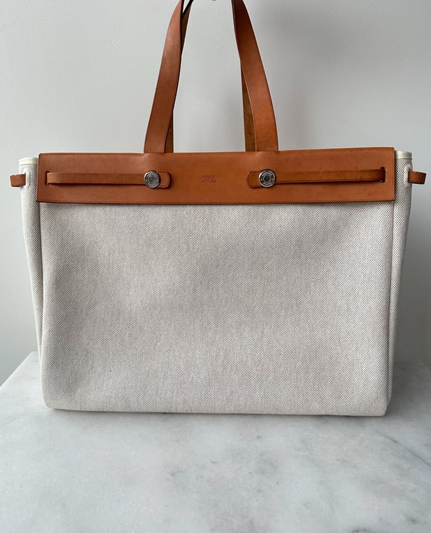 Beige stoftaske med brun læderkant og håndtag, placeret på en marmorflade foran en hvid væg.