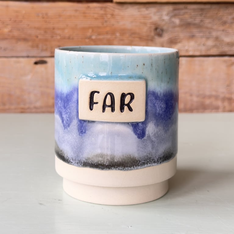 Krus med blå, lilla og sort glasur og en beige plade med teksten "FAR" foran, placeret på en hvid overflade med en træbaggrund.