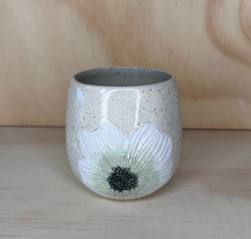 Keramikkrus med hvid blomst og grønne detaljer på lysebrun, prikket overflade.