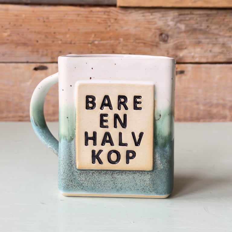 Firkantet keramikkrus med blågrøn glasur og teksten "BARE EN HALV KOP" på forsiden.