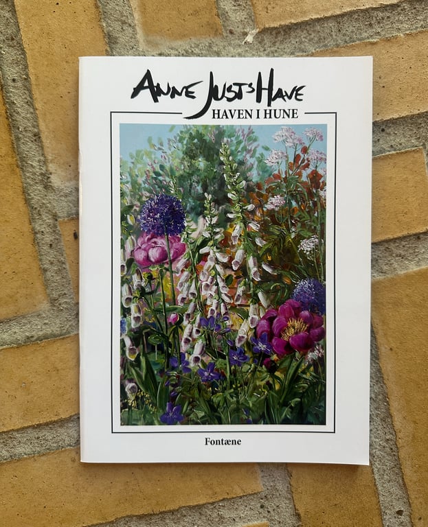 Billedet viser forsiden af bogen "Anne Just's Have - Haven i Hune" med et maleri af farverige blomster i forskellige nuancer af lilla, pink og grøn. Bogen ligger på et gulv af gule og grå fliser.