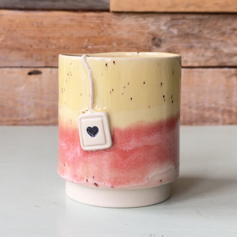 Keramisk kop med gul og pink glaseret overflade og en lille dekorativ tepose med hjerte hængende på ydersiden, stående på hvidt bord foran trævæg.