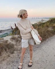 Kvinde iført lyse shorts og beige striktrøje står på en grussti ved kysten med havet og klitter i baggrunden ved solnedgang. (1)