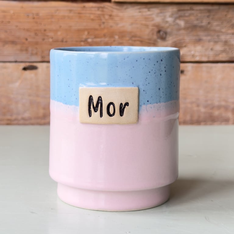 Keramikkrus med lyseblå og lyserød glasur og en beige plade med teksten "Mor" foran, placeret mod en træbaggrund.