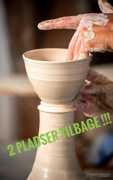 Hænder dækket af ler, der former en keramisk kop på en drejeskive med teksten "2 PLADSER TILBAGE !!!" i grøn på billedet.