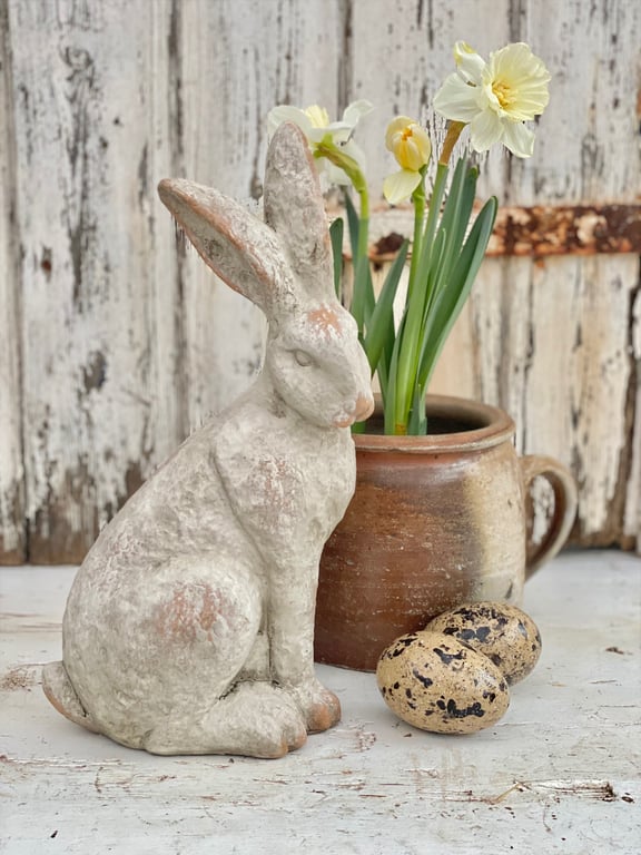 Dekorativ påskeharefigur i sten ved siden af en lerkrukke med påskeliljer og to plettede æg på en hvid, slidt træflade.