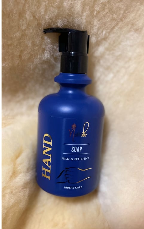 Flasken med håndsæbe i blå plast med sort pumpe, mærket "HAND SOAP - MILD & EFFICIENT - RIDERS CARE", liggende på blødt, lyst stof.