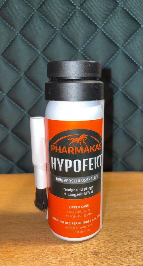 Flaske med Pharmakas Hypofekt lynlåsspray med børste, placeret på en træbordplade foran en mørk quiltet baggrund. (2)