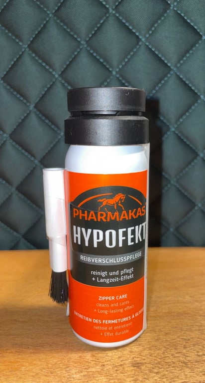 Flaske med Pharmakas Hypofekt lynlåsspray med børste, placeret på en træbordplade foran en mørk quiltet baggrund.
