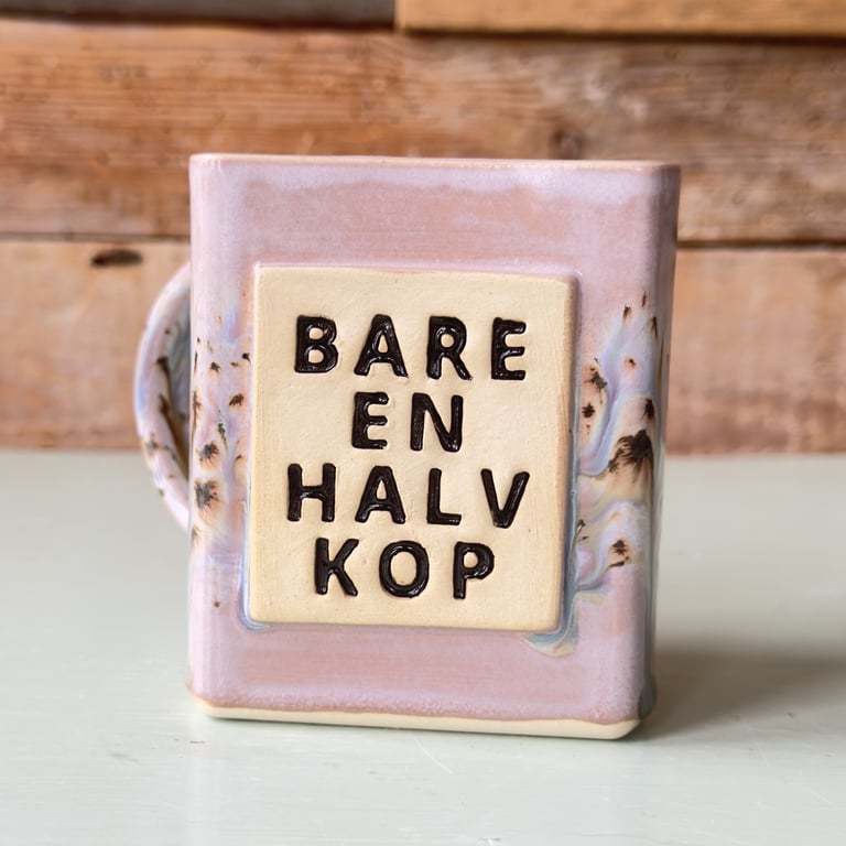 Firkantet keramisk kop med lys rosa glasur og tekst i sort "BARE EN HALV KOP" på forsiden.