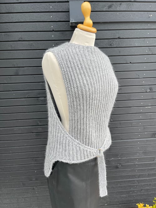 Grå strikket ærmeløs vest med bånd, vist på en mannequin foran en sort trævæg. (1)