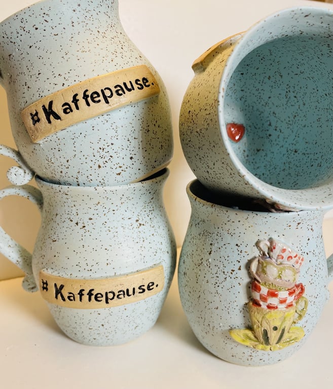 Billede af fire lyseblå keramikkrus med brune pletter. To krus har et påskrevet skilt med teksten "Kaffepause", og et krus har en dekorativ stak med farverige kopper.