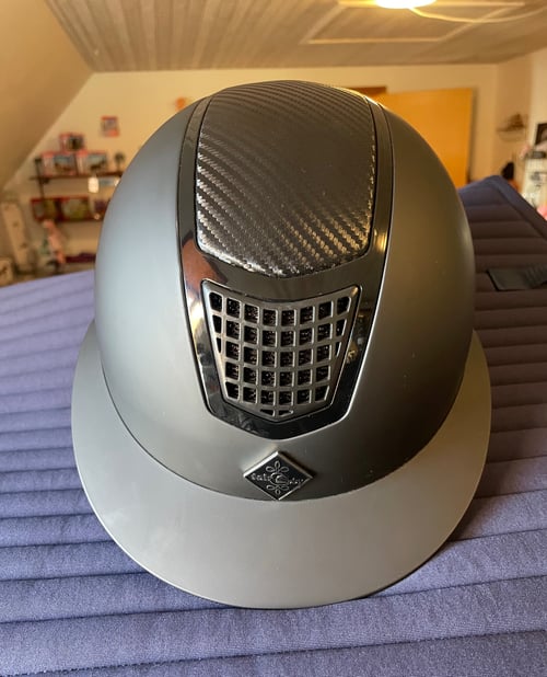 Sort og grå ridehjelm med ventilationsrist og logo, placeret på en lilla quiltet overflade indendørs. (1)