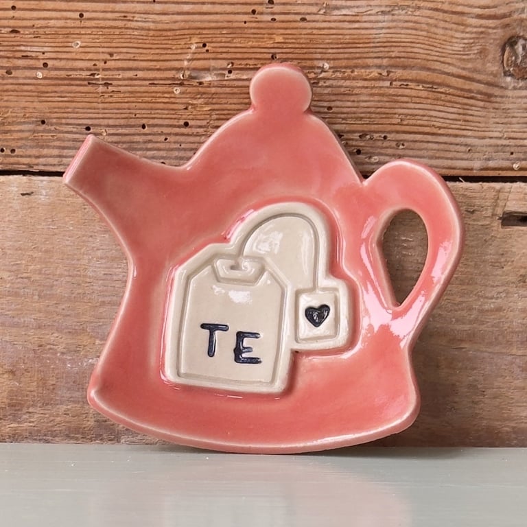 Rosa keramisk tekandeformet tallerken med udstikket tepose og teksten "TE" foran, stående på hvid overflade med træbaggrund.
