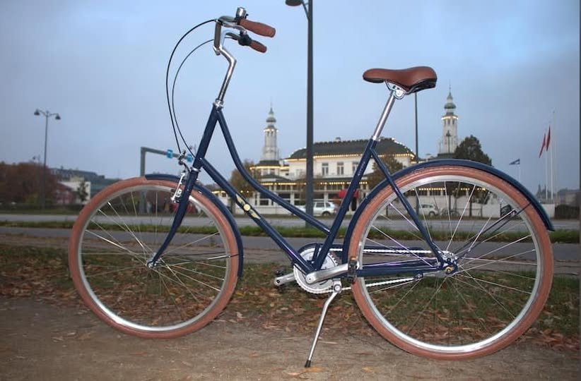 Billede af en klassisk blå cykel med brune dæk og saddel parkeret på en jordsti med en historisk bygning i baggrunden ved skumring.