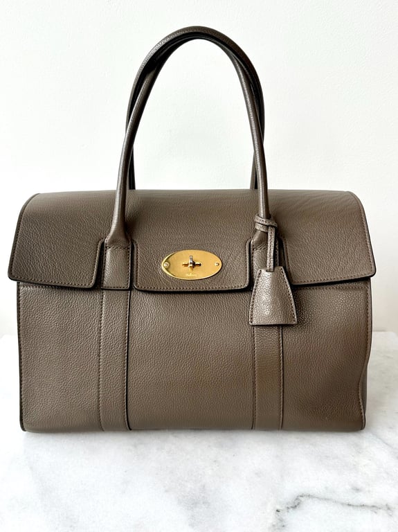 Brun læderhåndtaske med dobbelte håndtag og guldfarvet lås foran, placeret på hvid overflade.