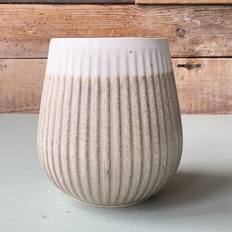 Keramisk krukke med lodrette riller i beige og hvid, placeret på en lys overflade foran en rustik trævæg.