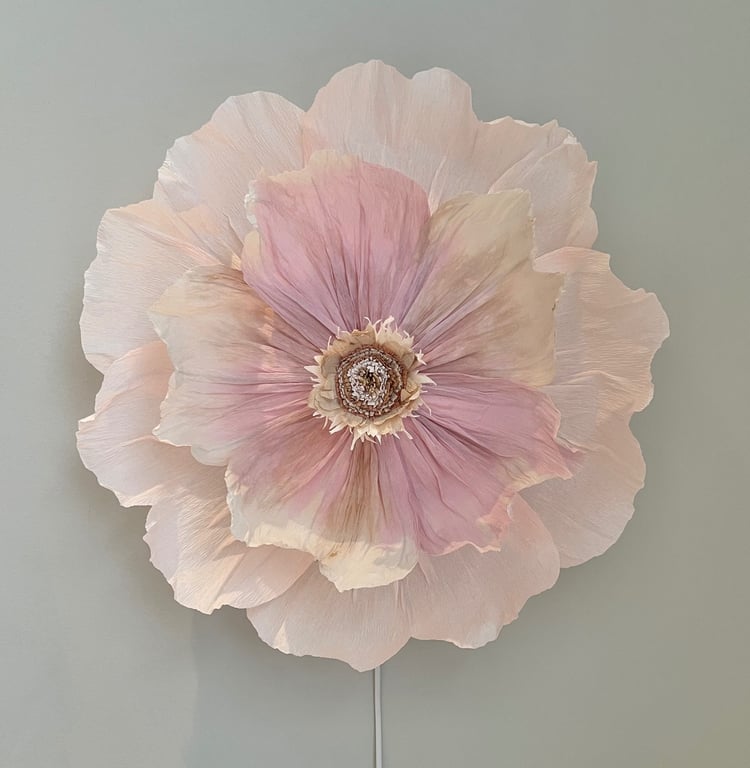 Stor kunstig blomst i sarte nuancer af lyserød og beige, monteret på en grå væg.