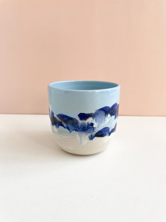 Lysblå keramisk kop med abstrakte mørkeblå og hvide mønstre, placeret på en hvid overflade mod en lyserød baggrund.