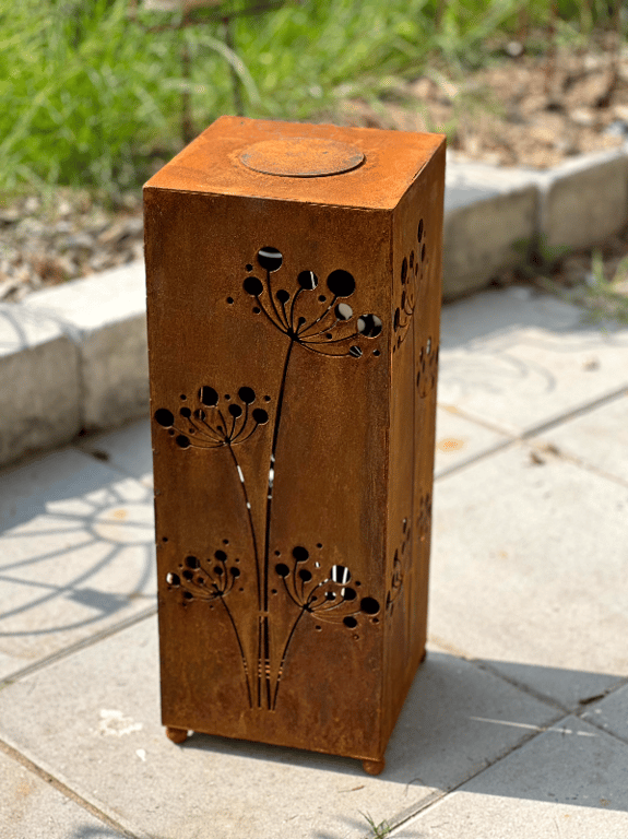 Rustik, firkantet metallanterne med blomsterudskæringer, placeret på flisebelagt gulv udendørs.