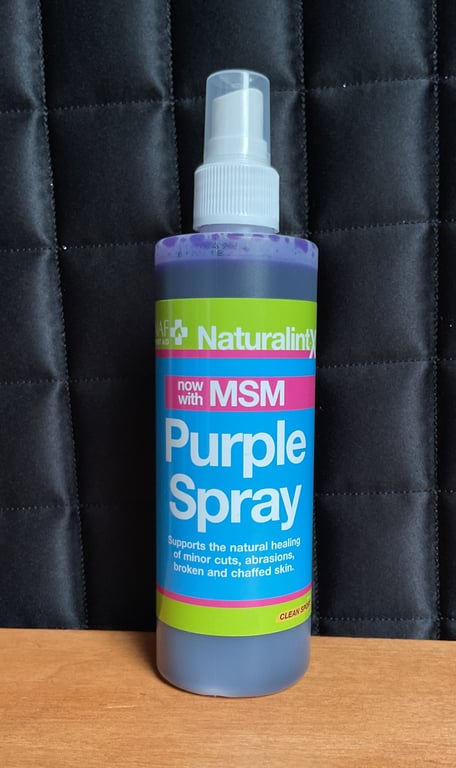 Flaske med Naturalints Purple Spray med MSM til heling af småsår, rifter og sprækket hud, placeret foran sort polstret baggrund.