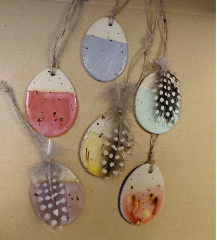 Fem påskeæg af keramik med forskellige farvede glasure og dekorative fjer, hængende i snore mod en beige baggrund.