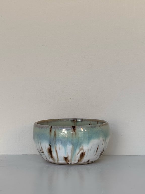 Keramisk skål med glasur i blå, hvid og brune nuancer stående på en grå overflade foran en beige væg.