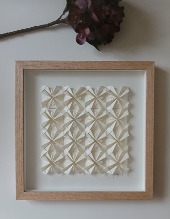 Indrammet kunstværk med tredimensionelle, foldede papirformer arrangeret i et geometrisk mønster, med en tørret blomstergren øverst. (2)