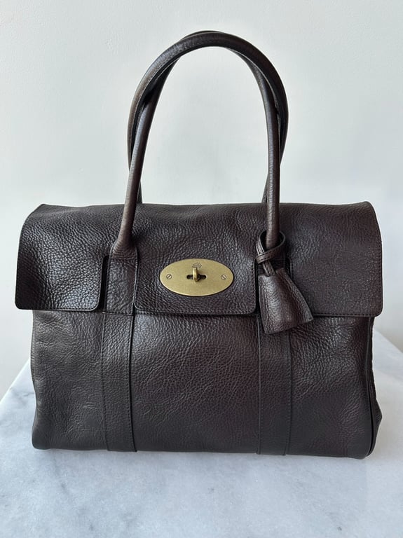 Brun lædertaske med korte håndtag og guldfarvet lås set forfra på marmorbord.