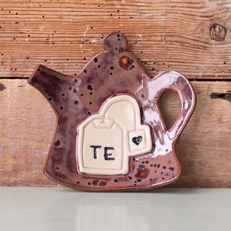 Keramisk teskeholder formet som en tekande med en relief af en tebrevsposer med teksten "TE" og et lille hjerte, placeret foran en rustik træbaggrund.