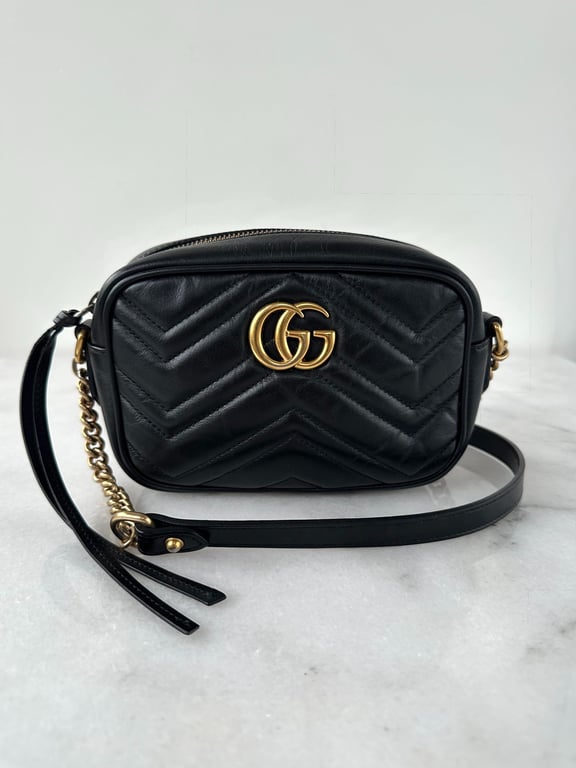 Sort lædertaske med guldfarvet GG-logo og kædedetalje på remmen, placeret på en hvid overflade.