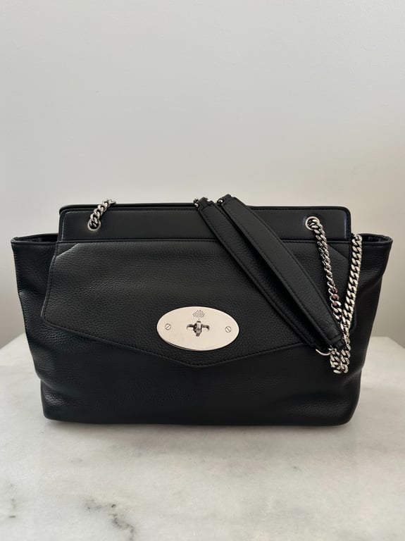 Sort lædertaske med sølvfarvet kæderem og oval spænde på fronten, placeret på en hvid marmorbordplade.