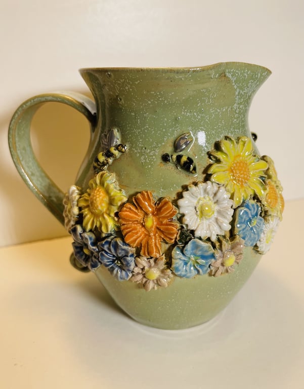 Grøn keramisk kande med relief af farverige blomster og bier.