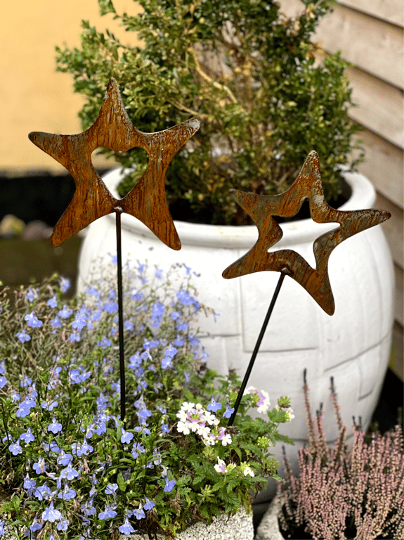 To rustne metalstjerner på pinde i en blomsterbed med lilla og hvide blomster, foran en stor hvid krukke med grøn plante.