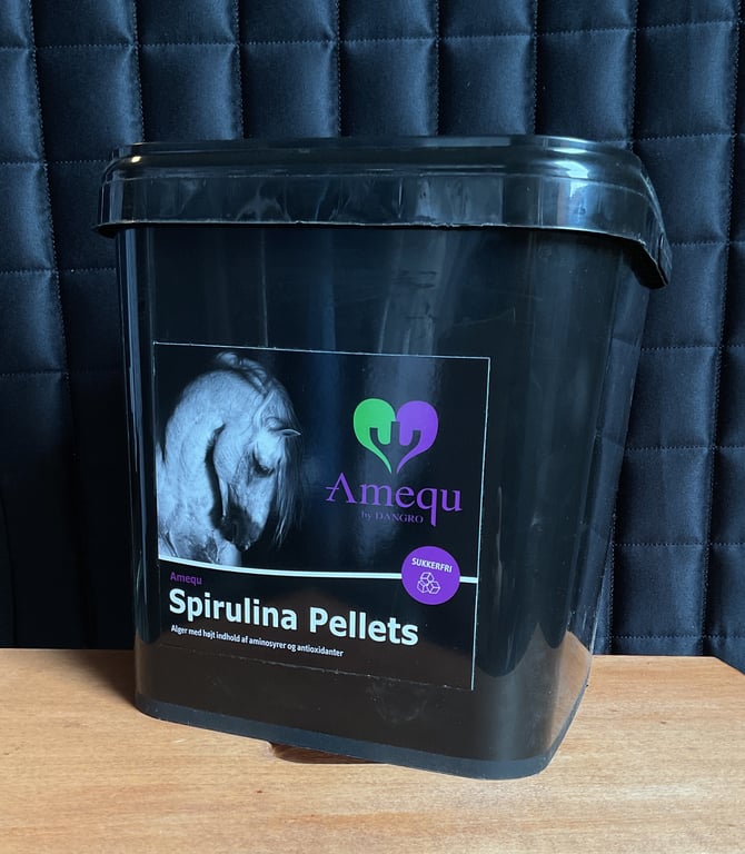 Sort spand med label til Amequ Spirulina Pellets, hestefoder med høj indhold af aminosyrer og antioxidanter, placeret på et træbord foran et mørkblåt quiltet stofbaggrund.