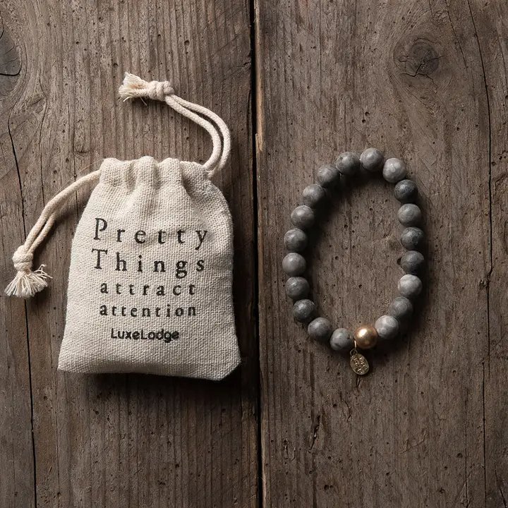 Smuk perlearmbånd med en guldfarvet perle og lille vedhæng ved siden af en stofpose med teksten "Pretty Things attract attention LuxeLodge" på en rustik træoverflade. (2)